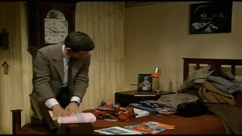A man in formal attire is seen folding clothes on a chair beside a bed, which is cluttered with various items, including magazines and other belongings. A clock hangs on the wall behind him.